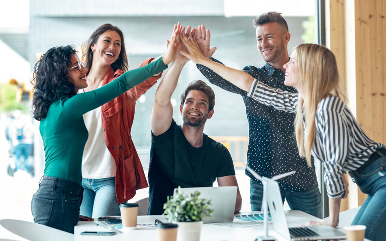 Eine Gruppe junger Berufstätiger am Arbeitstisch mit Laptop beim Feiern eines Erfolgs. Foto: nenetus - stock.adobe.com