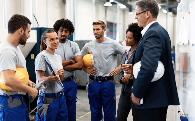 Diverses Produktionsteam mit Helmen und Arbeitshosen bei einer Besprechung. Foto: Drazen - stock.adobe.com