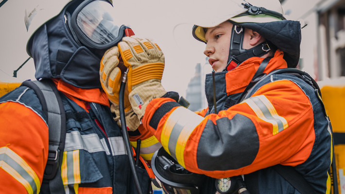 Ein Mann in Feuerwehrkleidung prüft bei einem Kollegen, ob die Atemschutzmaske richtig sitzt.