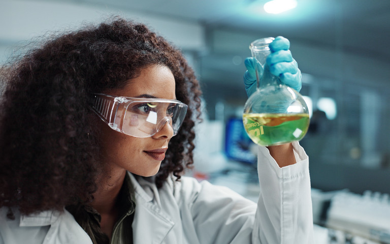 Frau mit Schutzbrille, die sich ein Reagenzglas mit grüner Flüssigkeit anschaut. Foto: Coetzee/peopleimages.com