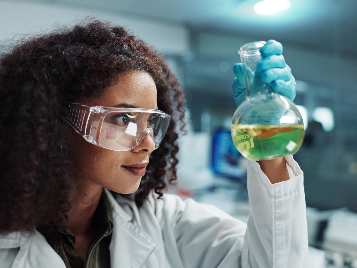 Frau mit Schutzbrille, die sich ein Reagenzglas mit grüner Flüssigkeit anschaut. Foto: Coetzee/peopleimages.com