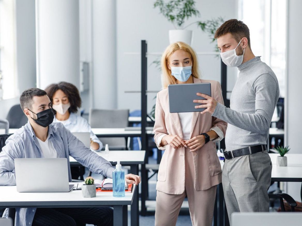 Teamwork in corporate company and social distance during coronavirus epidemic. Male manager in protective mask shows tablet to workers in interior.