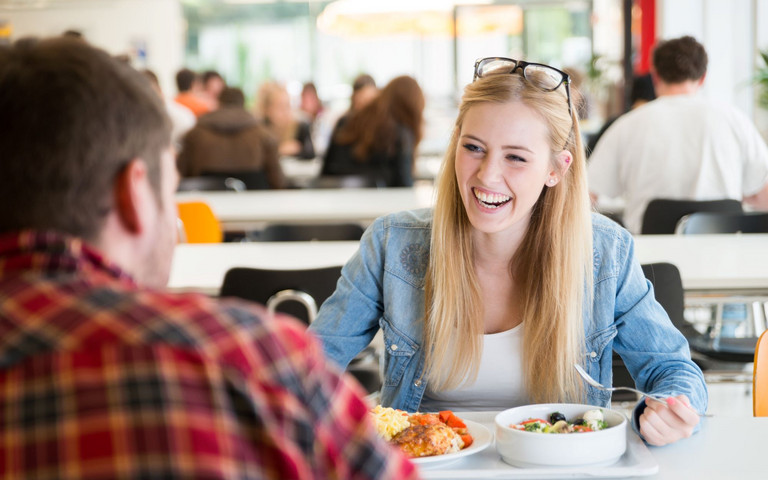 Junge Frau mit Gesprächspartner beim Essen in der Kantine. Foto: engel.ac - stock.adobe.com