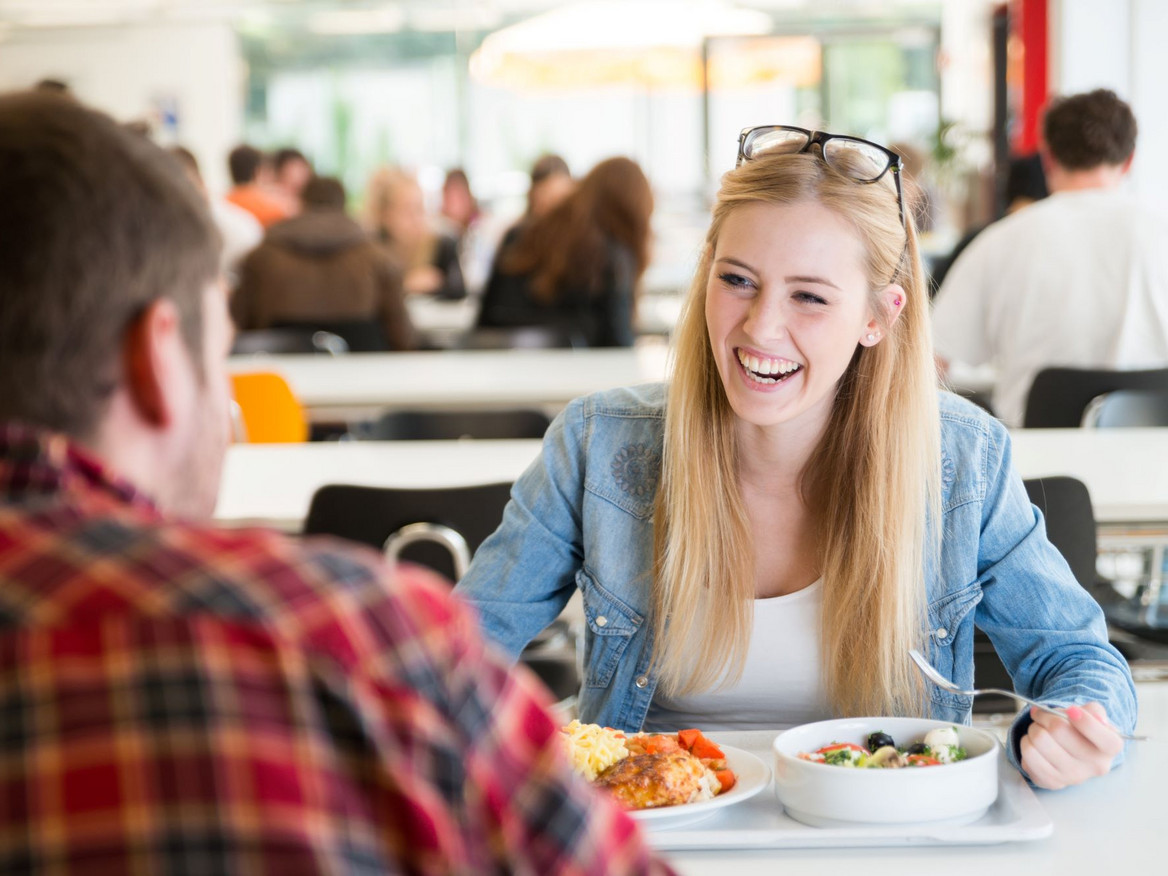 Junge Frau mit Gesprächspartner beim Essen in der Kantine. Foto: engel.ac - stock.adobe.com