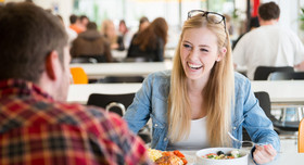 Junge Frau mit Gesprächspartner beim Essen in der Kantine. Foto: engel.ac - stock.adobe.com