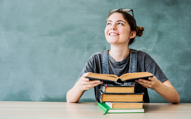 Frau sitzt an einem Tisch und liest ein Buch von einem Bücherstapel. Foto: sebra/stock.adobe.com 